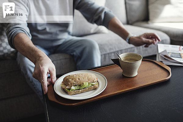 Mittelteil des älteren Mannes mit Sandwich und Tasse auf dem Sofa zu Hause sitzend
