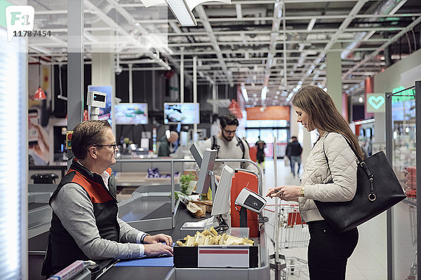 Reife Kassiererin betrachtet die Frau  die an der Kasse im Supermarkt durch Technologie zahlt.