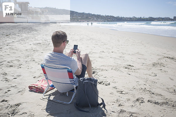 Rückansicht des Mannes  der ein Smartphone benutzt  während er auf einem Stuhl am Strand sitzt.