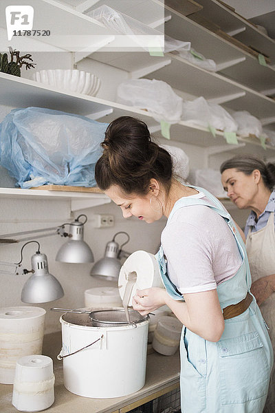 Senior Töpferin beim Anblick einer jungen Mitarbeiterin  die in der Werkstatt Ton aus der Vase in den Eimer gießt.
