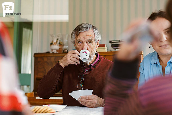 Senior Mann trinkt Kaffee  während er mit seiner Familie zu Hause Karten spielt.