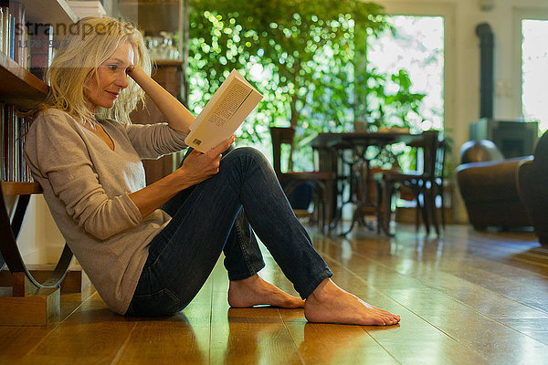 Reife Frau sitzt zu Hause auf dem Boden und liest Buch.