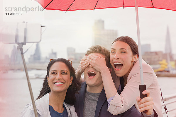 Verspielter Freund Touristen mit Regenschirm nimmt Selfie mit Fotohandy Selfie Stick auf Brücke  London  UK