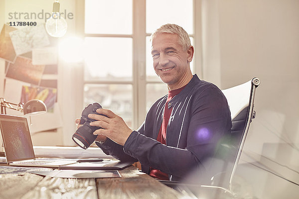 Portrait lächelnder  selbstbewusster Fotograf mit Digitalkamera bei der Arbeit am Laptop im sonnigen Büro