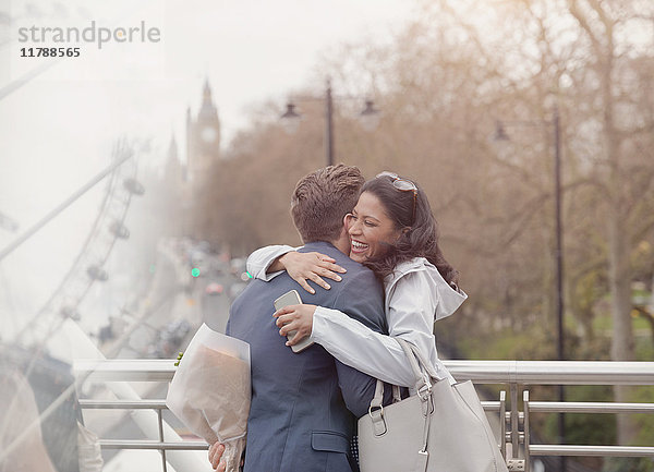 Lächelndes Paar umarmend  Freund überraschende Freundin mit Blumen auf urbaner Brücke  London  UK