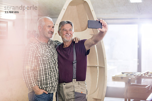 Stolze  lächelnde Schreiner mit Fotohandy  die Selfie neben dem Holzboot in die Werkstatt bringen.