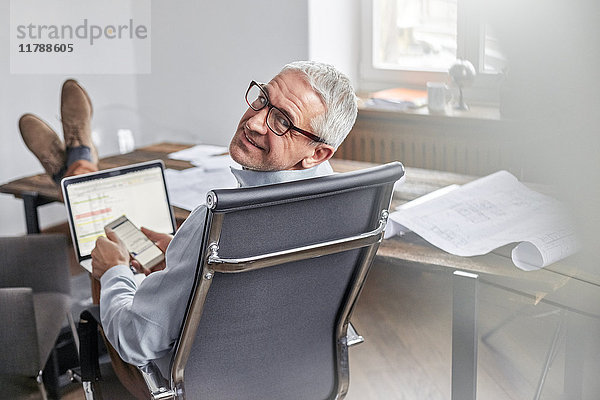 Portrait lächelnder  selbstbewusster Geschäftsmann mit Handy und Laptop im Büro