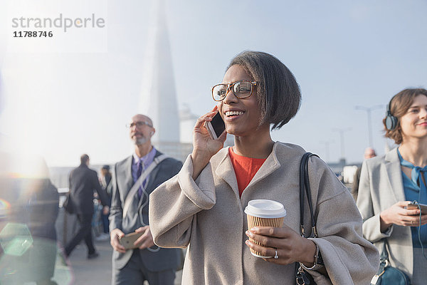 Lächelnde Geschäftsfrau trinkt Kaffee und telefoniert auf der sonnigen  belebten Fußgängerbrücke der Stadt