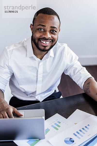 Porträt eines lächelnden Mannes  der Daten analysiert und den Laptop am Schreibtisch im Home-Office benutzt.