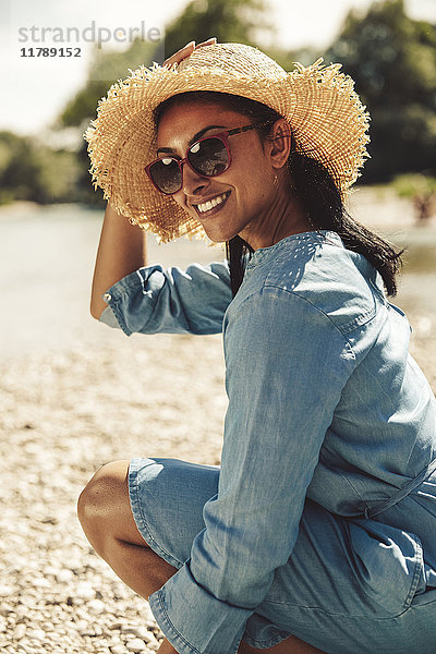 Porträt einer glücklichen Frau mit Strohhut und Sonnenbrille am Strand