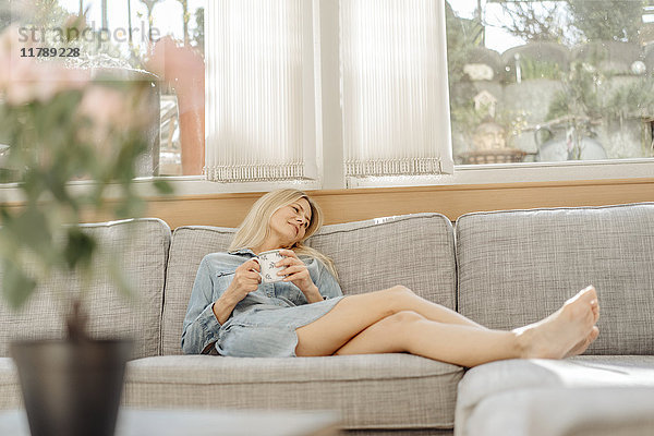 Frau zu Hause auf der Couch liegend mit Tasse Kaffee