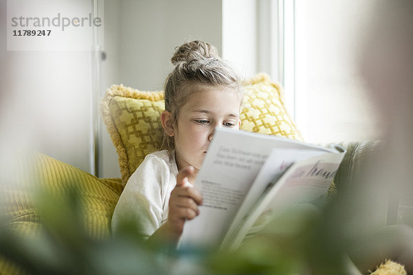 Porträt eines entspannten kleinen Mädchens  das auf einem Sessel sitzt und die Zeitschrift liest.