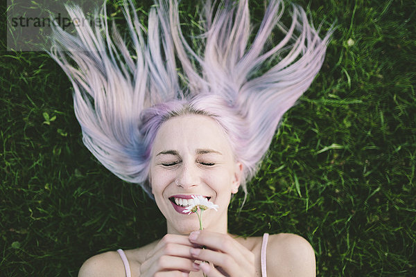 Porträt einer lachenden Frau  die mit geschlossenen Augen auf Gras liegt und Gänseblümchen hält.