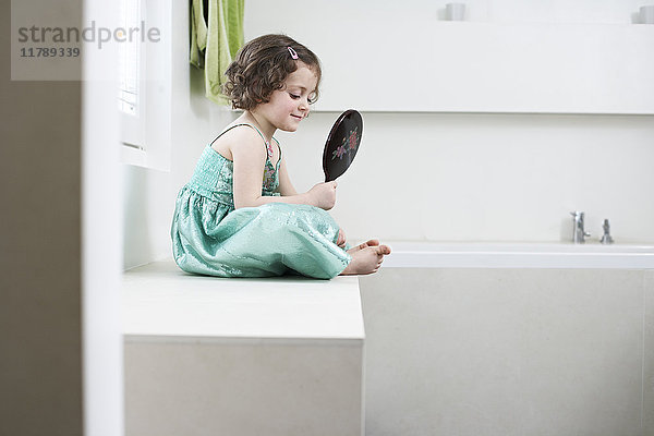 Lächelndes kleines Mädchen  das sich in einem Handspiegel im Badezimmer sieht.