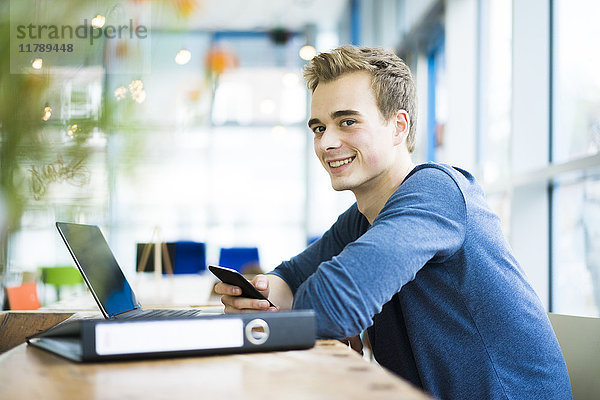 Porträt eines lächelnden Schülers mit Latop  Handy und Akte im Café