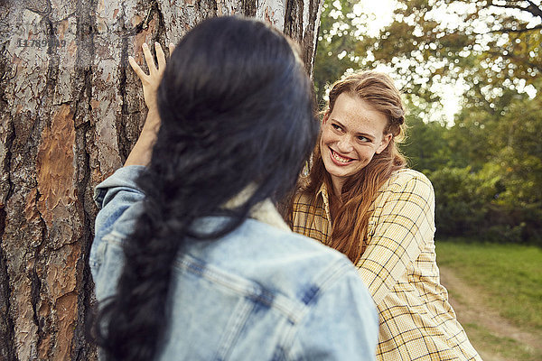 Porträt einer rothaarigen jungen Frau  die sich mit ihrem Freund im Vordergrund an den Baumstamm lehnt.