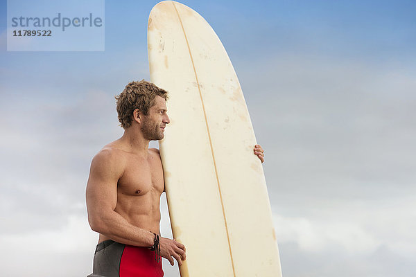 Mann steht am Strand mit Surfbrett und genießt die Aussicht