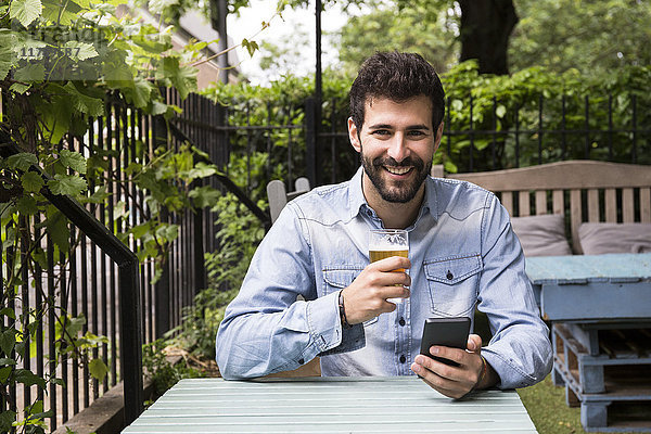 Porträt eines lächelnden jungen Mannes mit Handy und einem Glas Bier im Garten