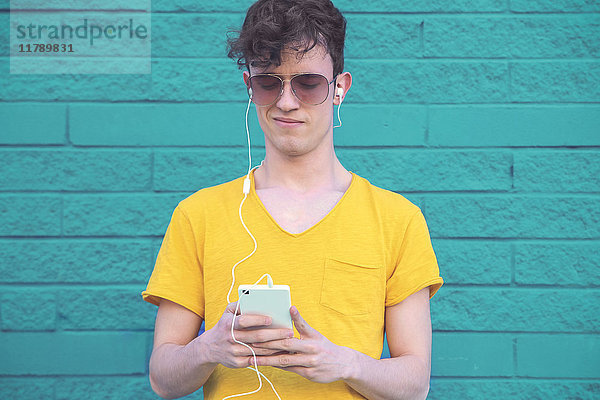Portrait eines jungen Mannes beim Musikhören mit Smartphone und Kopfhörer vor blauer Ziegelwand