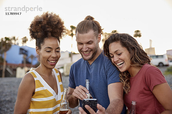 Drei lachende Freunde mit Bierflaschen beim Blick auf das Smartphone am Strand