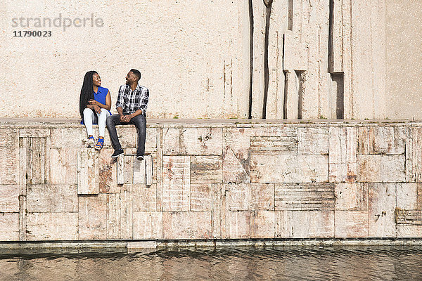 Sprechendes junges Paar auf einer Wand am Wasser sitzend