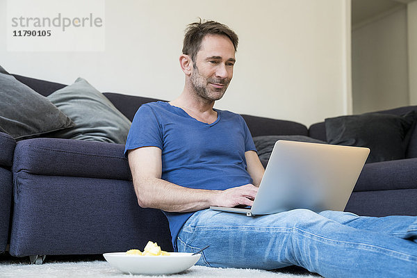 Porträt eines lächelnden Mannes  der im Wohnzimmer mit dem Laptop auf dem Boden sitzt.