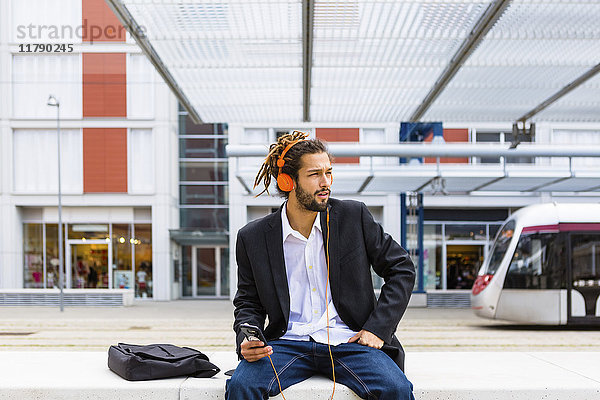 Junger Geschäftsmann mit Dreadlocks beim Musikhören mit Kopfhörer und Handy am Bahnhof