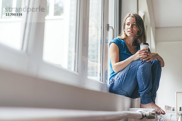 Lächelnde junge Frau mit Kaffee zum Mitnehmen am Fenster sitzend