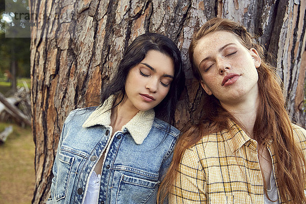 Porträt zweier junger Frauen  die sich mit geschlossenen Augen an den Baumstamm lehnen.
