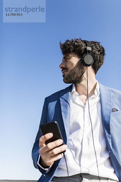 Junger Geschäftsmann mit Smartphone mit Kopfhörer unter blauem Himmel