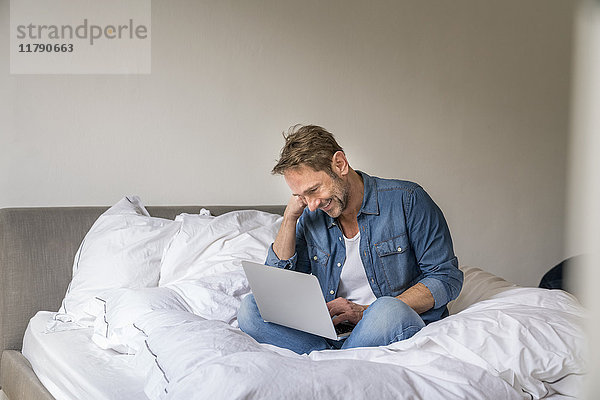 Lächelnder reifer Mann auf seinem Bett sitzend mit Laptop