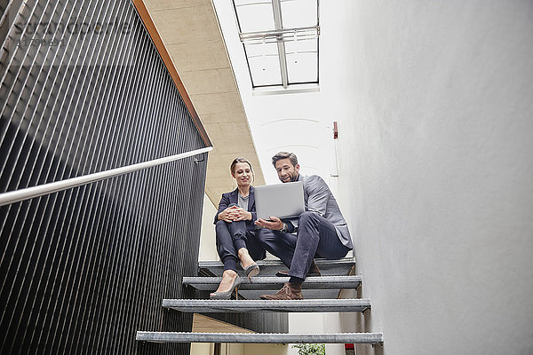Geschäftsmann und Geschäftsfrau sitzen auf einer Treppe im Büro und teilen sich einen Laptop.