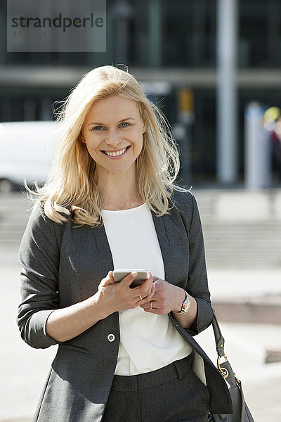 Portrait der lächelnden blonden Geschäftsfrau mit Handy und Handtasche