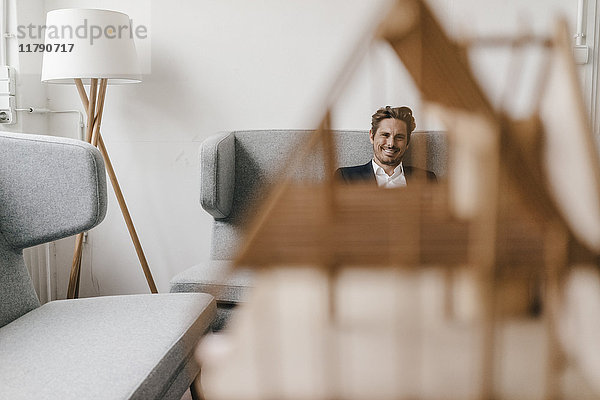 Lächelnder Mann auf der Couch mit Architekturmodell im Vordergrund