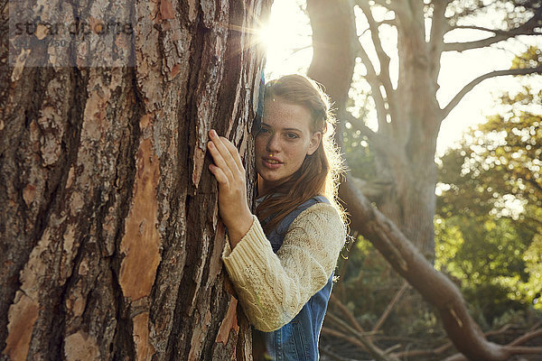 Porträt einer jungen Frau  die im Gegenlicht den Baum umarmt