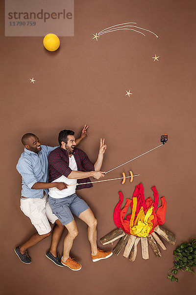 Schwules Paar brät Würstchen am Lagerfeuer und nimmt sich einen Selfie.