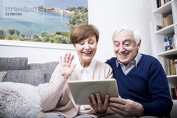 Seniorenpaar sitzt auf der Couch und macht einen Videoanruf auf ihrem digitalen Tablett.