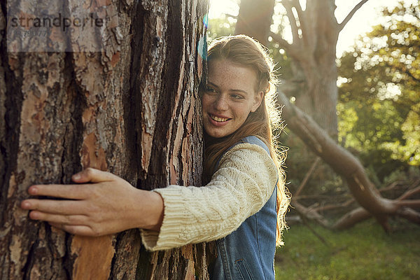 Porträt einer lächelnden jungen Frau  die den Baumstamm im Gegenlicht umarmt.