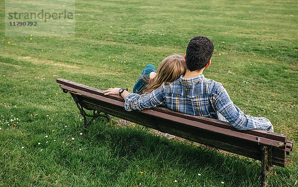 Rückansicht eines jungen Paares  das auf einer Bank auf einer Wiese sitzt.
