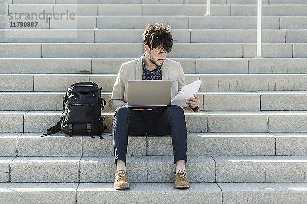 Nachdenklicher Mann mit Laptop  der auf einer Treppe sitzt und Dokumente überprüft.