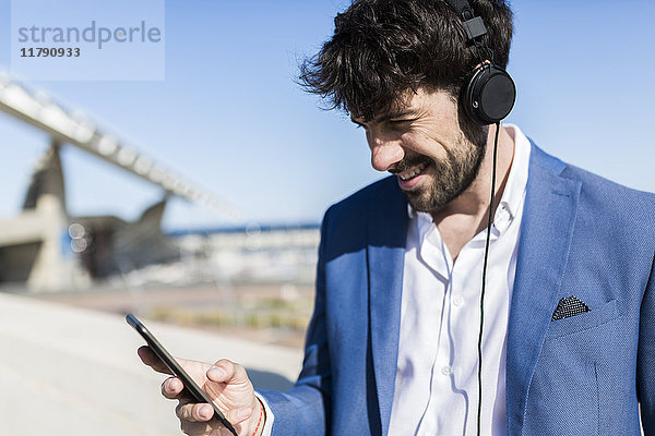 Junger Geschäftsmann mit Kopfhörer und Blick auf das Smartphone im Freien