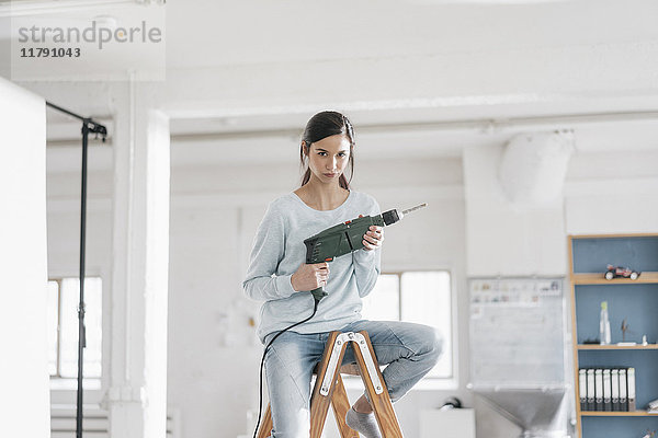 Junge Frau in ihrer neuen Wohnung sitzt auf einer Leiter und hält eine elektrische Bohrmaschine.