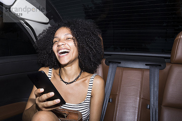 Porträt einer lachenden Frau mit Smartphone auf den Rücksitzen eines Autos
