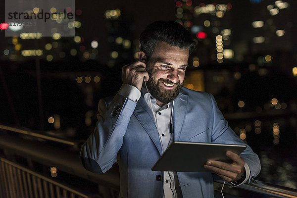 Lächelnder junger Mann mit Tablett und Kopfhörer auf Stadtbrücke bei Nacht