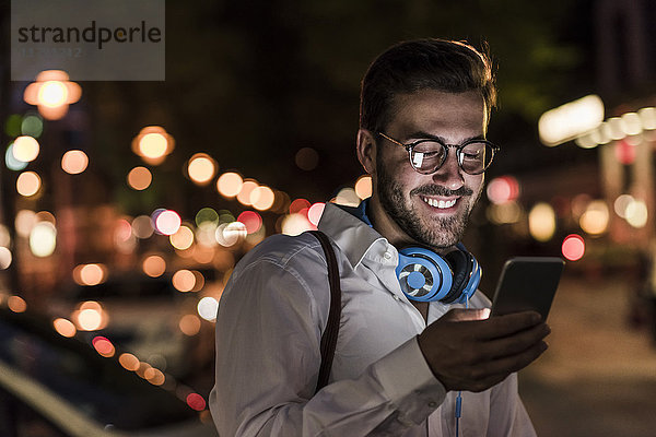 Lächelnder junger Mann in der Stadt  der nachts das Handy überprüft.