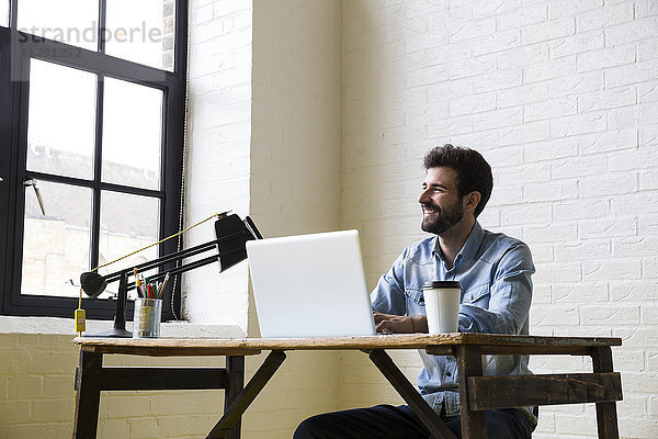 Lächelnder Mann sitzt am Schreibtisch und arbeitet mit seinem Laptop.