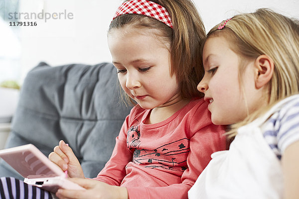 Zwei kleine Mädchen sitzen nebeneinander auf der Couch und schauen aufs Handy.