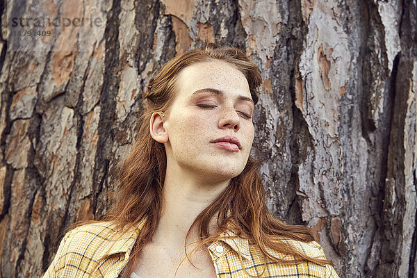 Porträt einer rothaarigen jungen Frau  die sich mit geschlossenen Augen an den Baumstamm lehnt.