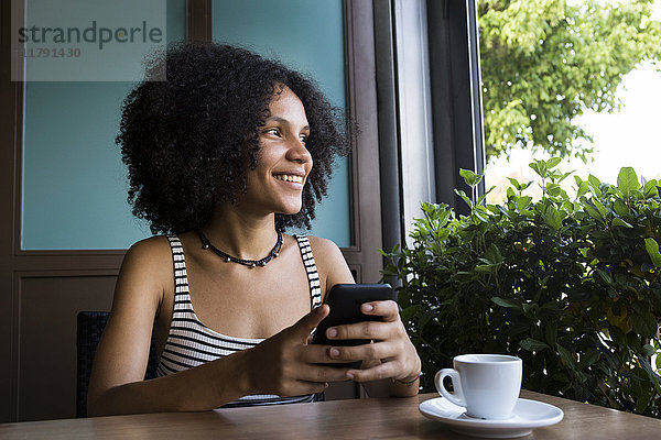 Junge Frau mit Smartphone sitzt in einem Café und schaut aus dem Fenster.