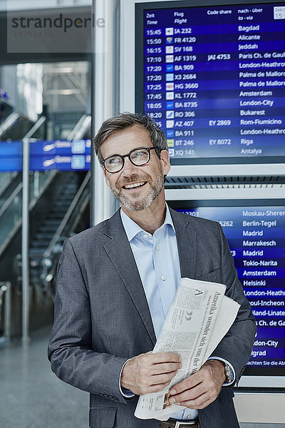 Lächelnder Geschäftsmann mit Zeitung zum Flugplan am Flughafen